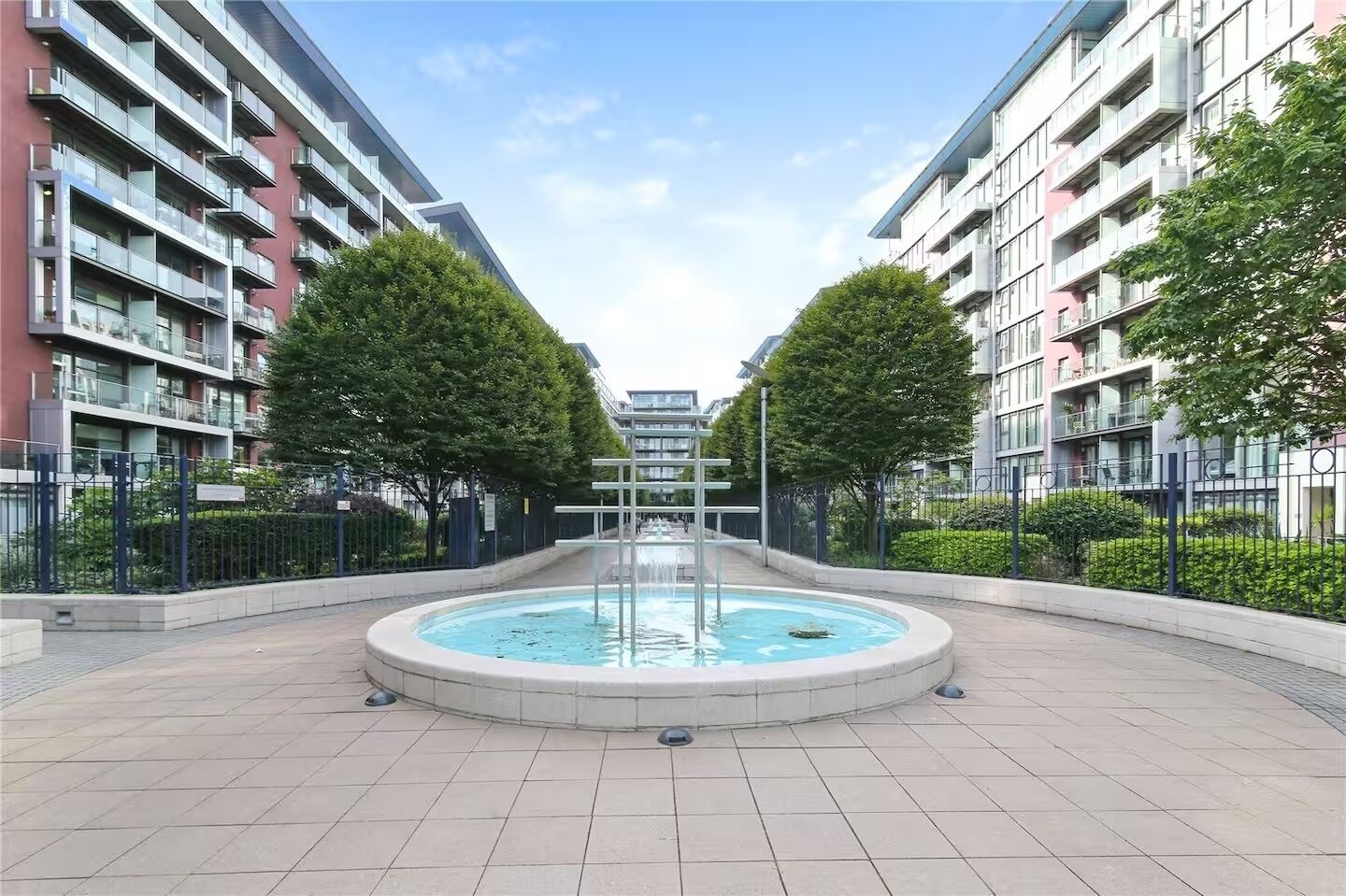 Ein moderner Apartmentkomplex mit einem zentralen Brunnen, umgeben von Bäumen und einem gepflasterten Weg.