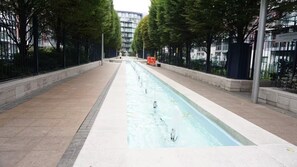 Ein langer, schmaler Wasserlauf in einer begrünten Stadtlandschaft, mit Bäumen und einem Gebäude im Hintergrund.