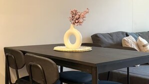 Uma sala de jantar com mesa, cadeiras, um vaso com flores e um sofá ao fundo.