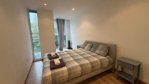 Quarto com cama, mesa de cabeceira, janela com cortinas e vista para o verde lá fora.