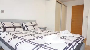 Ein ordentlich bezogenes Bett mit karierter Bettdecke, ein Nachttisch mit Lampe und eine Holztür im Hintergrund.