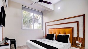 A bedroom with a bed, bedside tables, a desk, and a window with a view of palm trees. - Super Deluxe Room Kris Resort (Calangute)