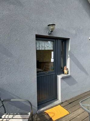 Ein Gebäude mit strukturierter Fassade, einer dunklen Tür mit Fenster, einer kleinen Ablage mit einer Figur und einem Metallstuhl.
