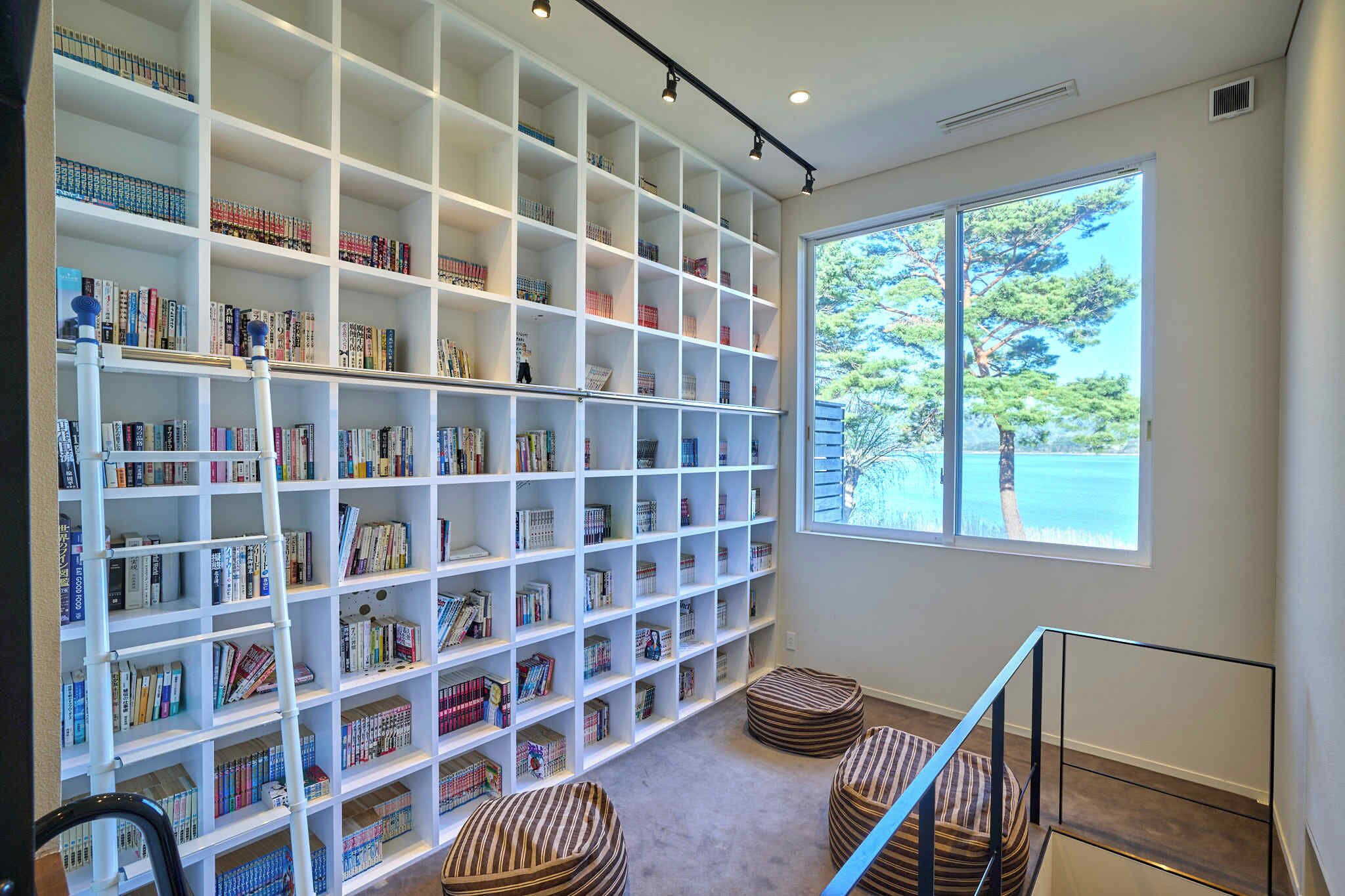 A spacious room with a large bookshelf filled with books, a window with a view of trees, and a staircase with a railing.