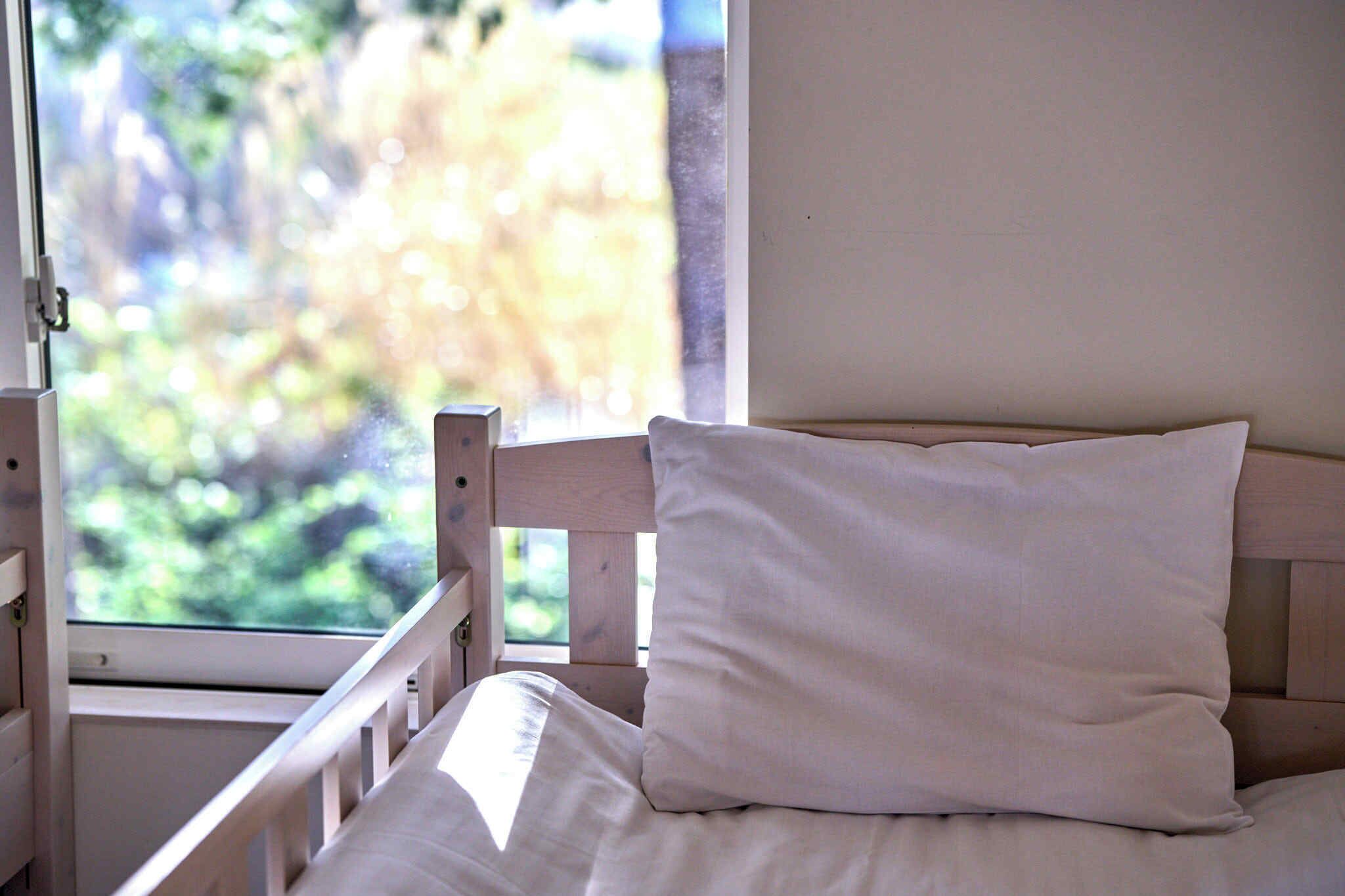 Un letto con cuscino bianco e testiera in legno, una finestra con vista sul verde e una parete bianca.
