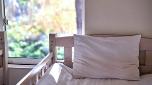 Una cama con almohada blanca y cabecera de madera, una ventana con vistas a follaje, y una pared blanca.