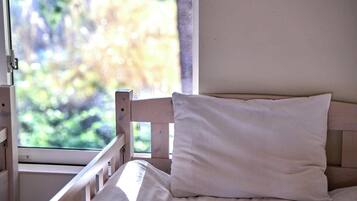 Un letto con cuscino bianco e testiera in legno, una finestra con vista sul verde e una parete bianca.