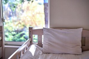 Una cama con almohada blanca y cabecera de madera, una ventana con vistas a follaje, y una pared blanca.