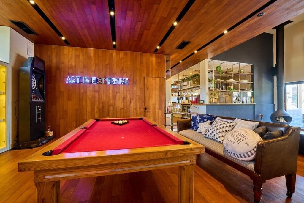 A pool table in a room with wooden ceiling and walls, a neon sign, a sofa with cushions, and a bar area in the background.