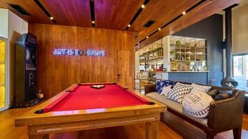 A pool table in a room with wooden ceiling and walls, a neon sign, a sofa with cushions, and a bar area in the background.