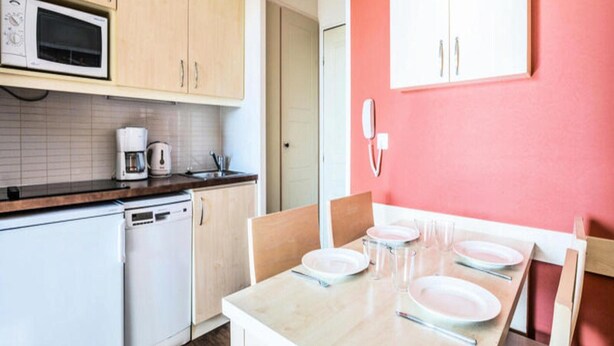 A kitchen with wooden cabinets, a microwave, a coffee maker, and a dining table set with plates and glasses. - Residence The Parks of Grimaud (Grimaud)