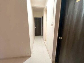 A narrow hallway with a dark door at the end, white walls, and a light switch on the wall.