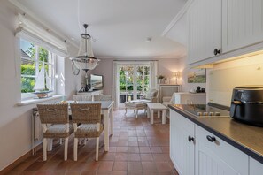 A kitchen with white cabinets, a dining area with wicker chairs, a chandelier, and a view through a door. - Wencke - Westerland (Westerland)