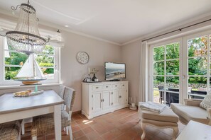 A room with a dining table, chairs, a white cabinet, a television, and a large window with a view of greenery. - Wencke - Westerland (Westerland)
