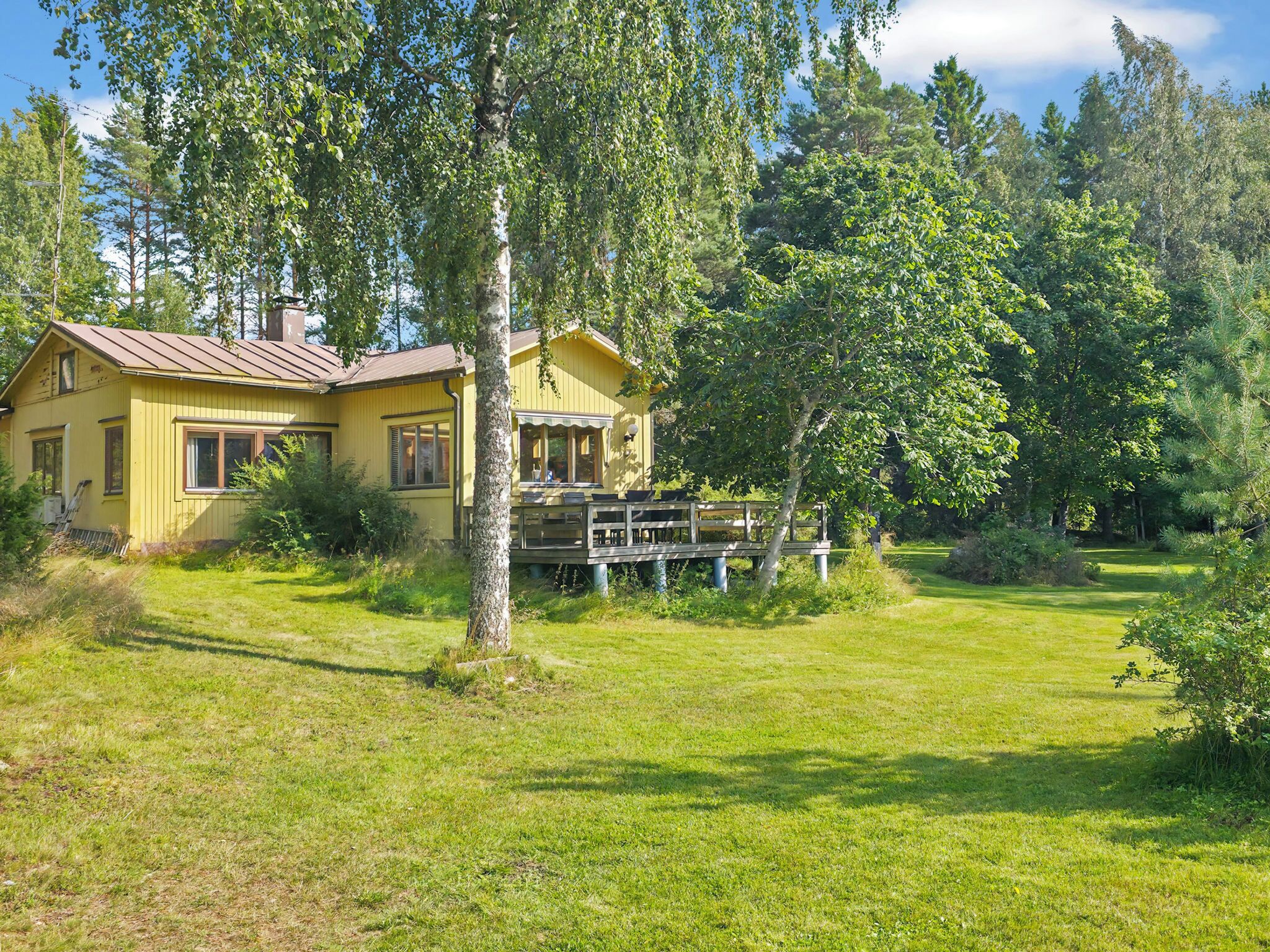 A yellow house with a porch and a deck, surrounded by a grassy yard and trees.