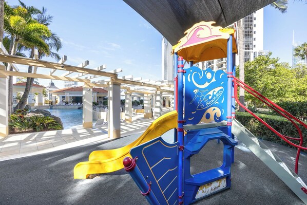 Estructura de juegos infantiles con tobogán, cerca de una piscina y palmeras, con edificios altos al fondo.