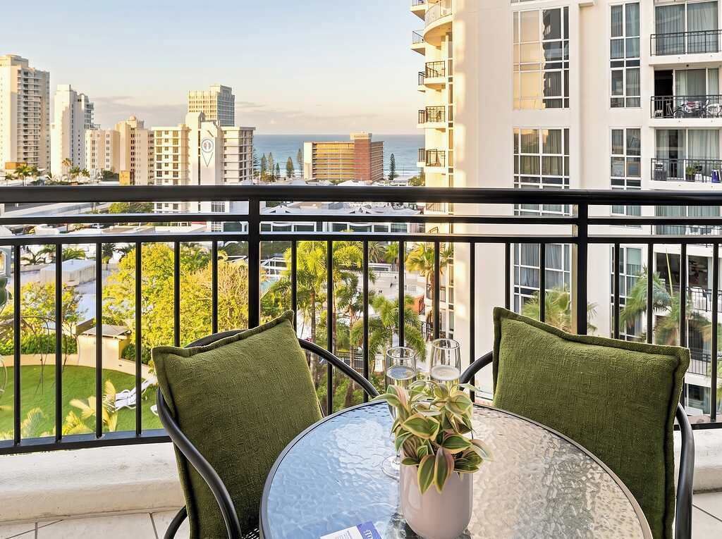 Un balcon avec une table et des chaises, donnant sur un paysage urbain avec des immeubles et une vue sur l’océan.