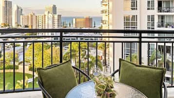 Un balcon avec une table et des chaises, donnant sur un paysage urbain avec des immeubles et une vue sur l’océan.