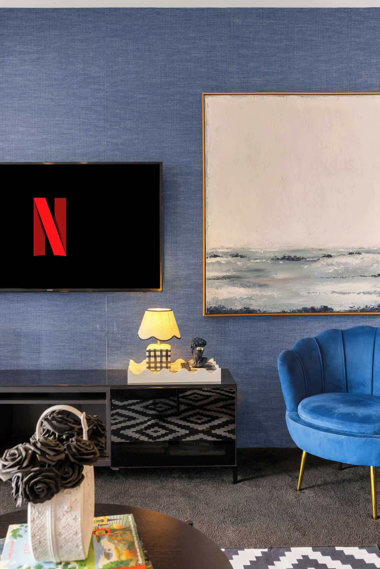A modern living room with a flat-screen TV, a blue armchair, a lamp, a sculpture, and a painting on the wall.