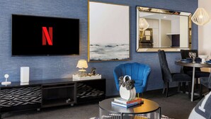 A modern living room with a TV displaying a red 'N', a large framed artwork, a blue armchair, a coffee table with books, and a dining area with chairs and a table. - Sunrise 19 at Circle on Cavill | 2BR Ocean View (Surfers Paradise)