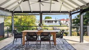 Eine überdachte Terrasse mit Tisch und Stühlen, einem Deckenventilator und Blick auf ein Haus und Grünflächen.