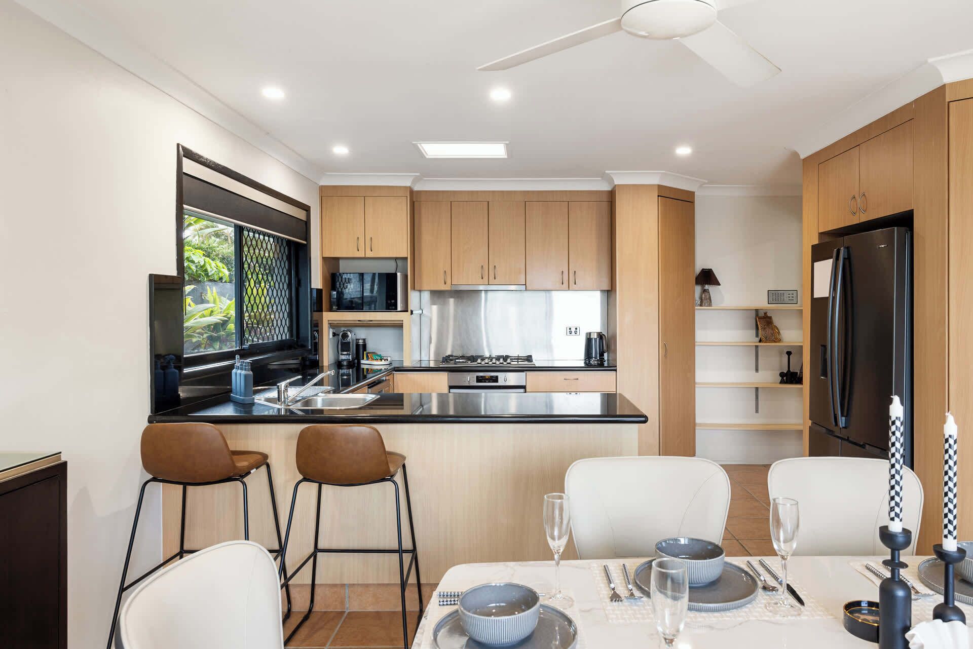 A modern kitchen with wooden cabinets, a stainless steel refrigerator, and a dining area with white chairs and a table set for two.