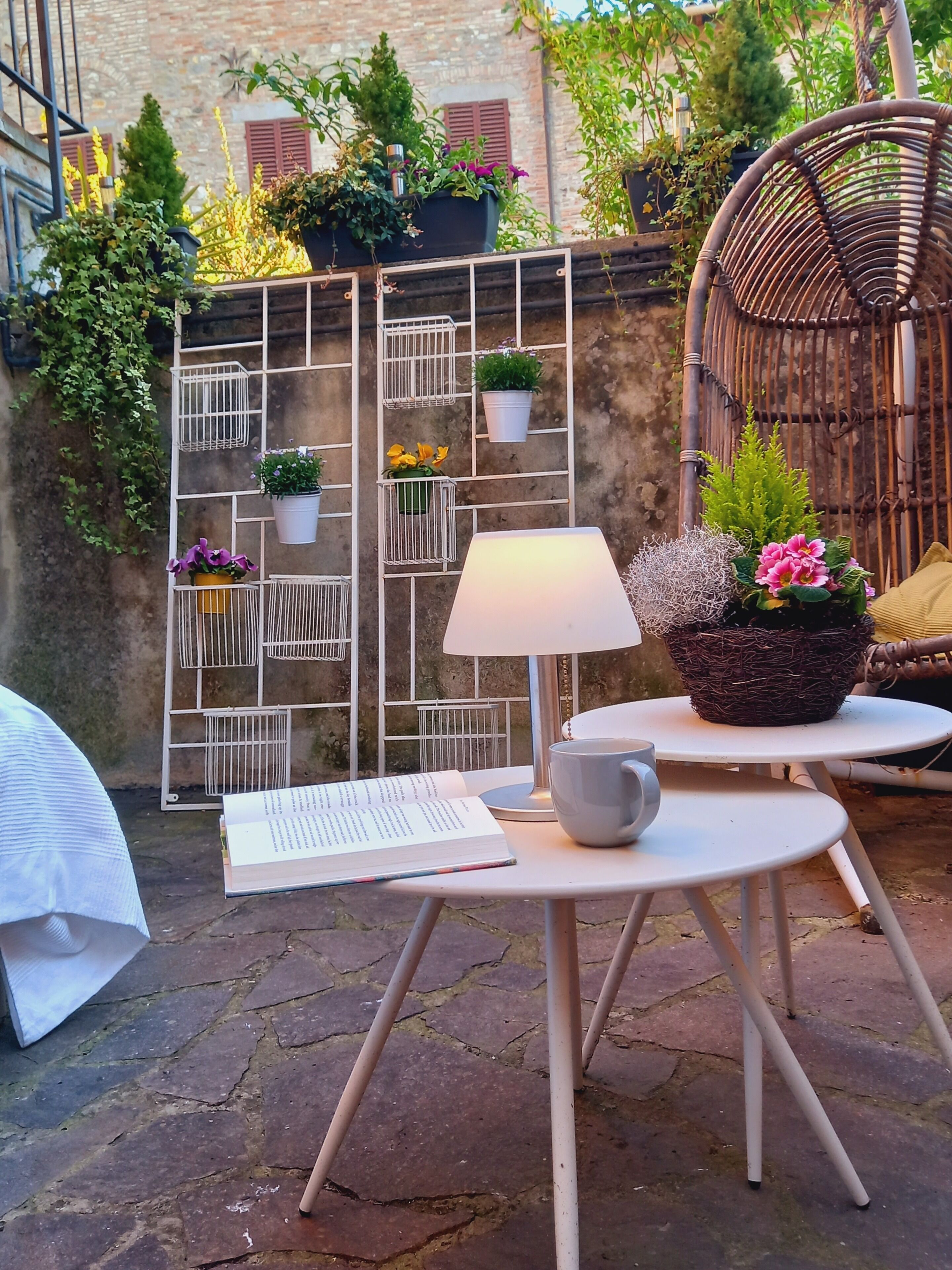 Un área de estar al aire libre con una mesa, una silla y una lámpara. Hay plantas en macetas y una silla de mimbre al fondo.