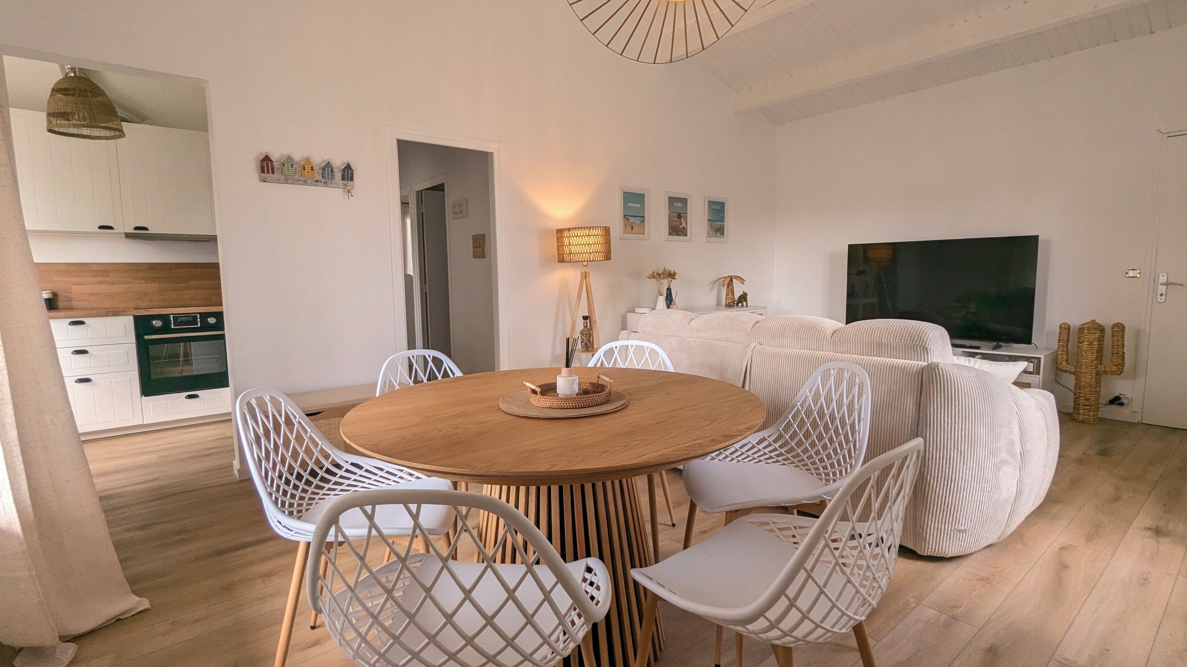 Ein modernes Wohnzimmer mit einem runden Holzes Esstisch, weißen Korbstühlen, einem Flachbildfernseher und einer Küche im Hintergrund.