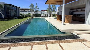 Uma área de piscina moderna ao ar livre, com piscina azul cristalina, ladrilhos de pedra e um espaço de estar coberto com cozinha integrada.
