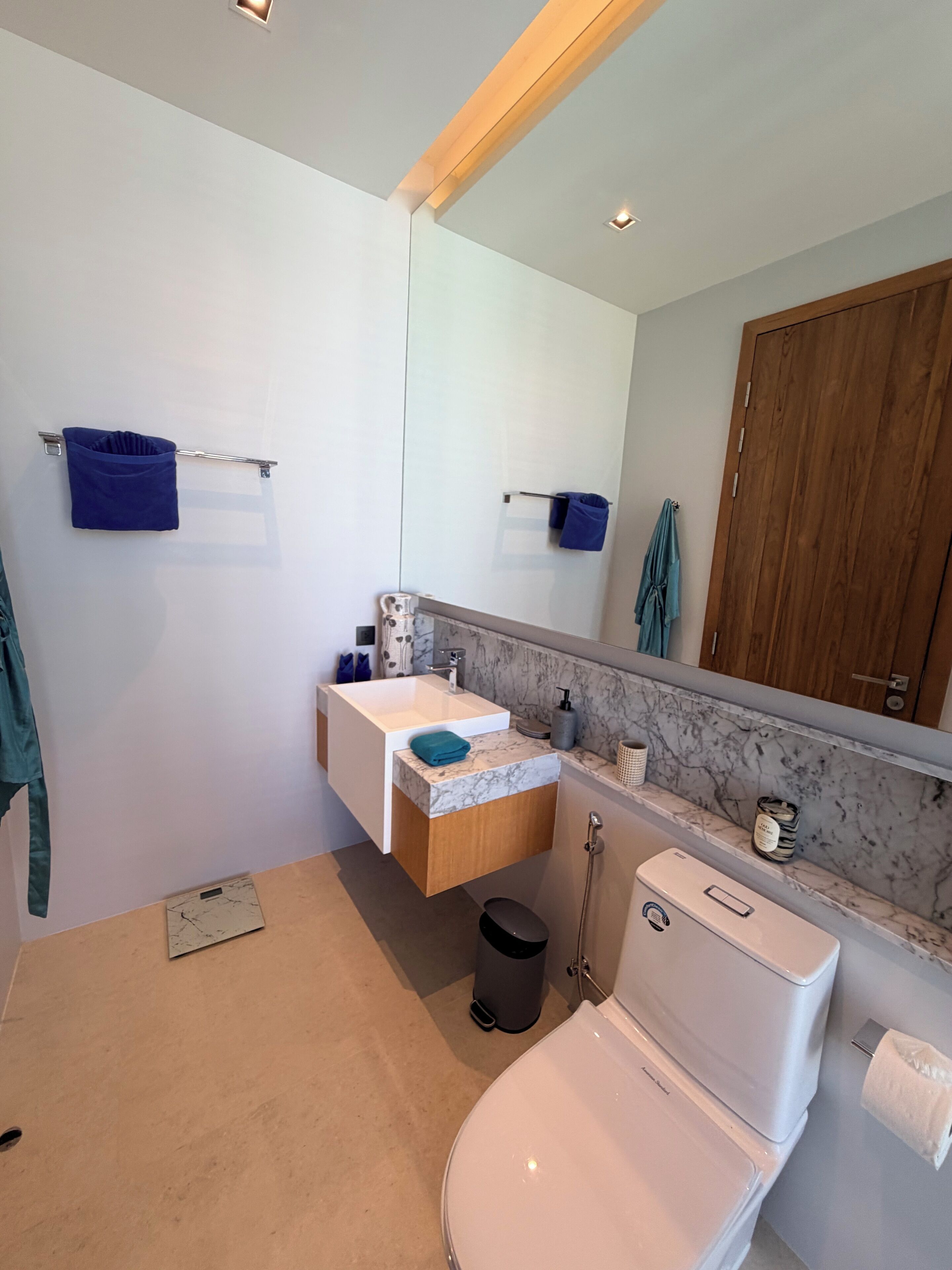 A modern bathroom with a white toilet, a sink with a marble countertop, a wooden cabinet, and a large mirror.