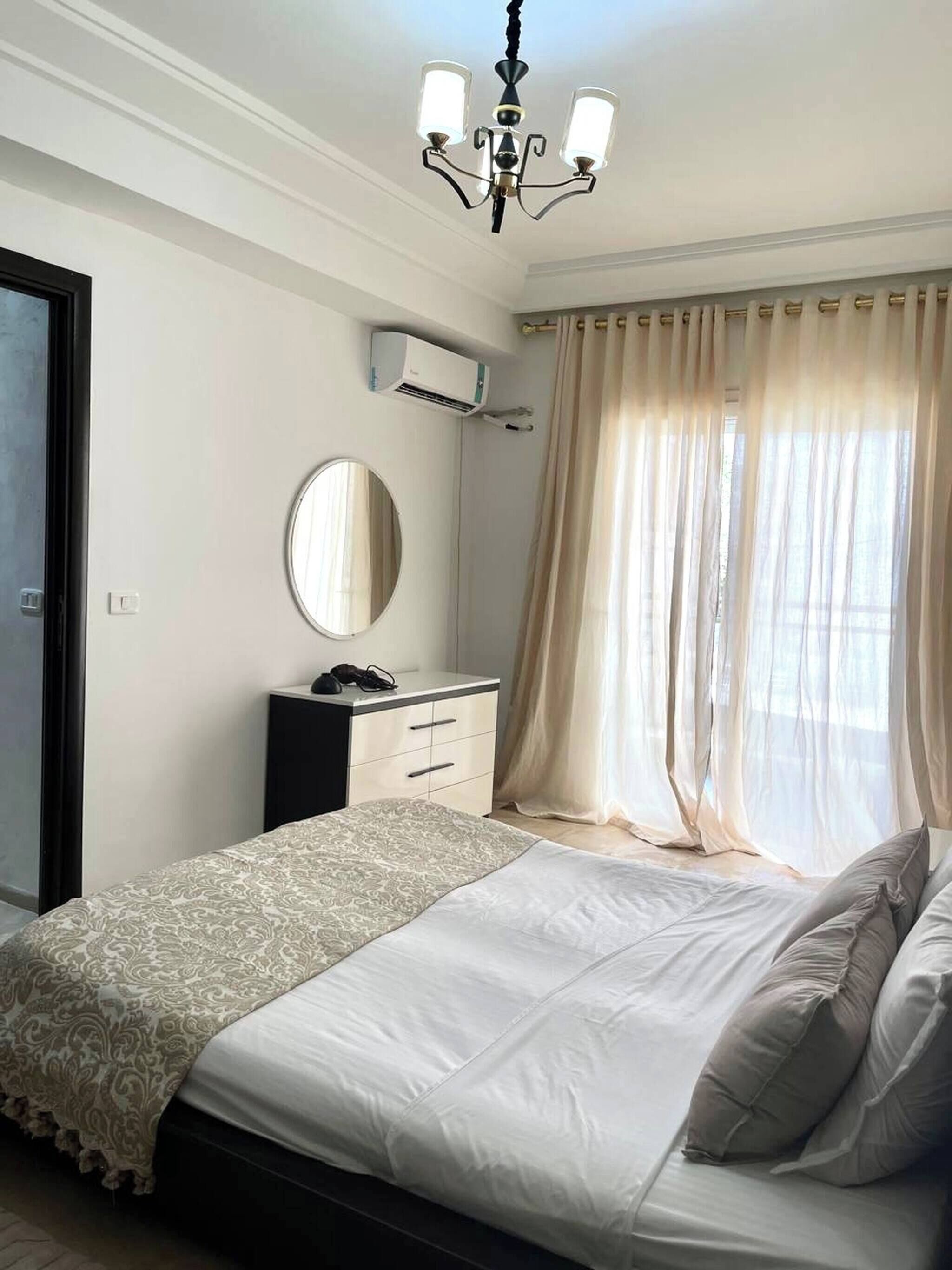 Une chambre à coucher comprenant un lit, une commode, un miroir, un appareil de climatisation et une fenêtre avec des rideaux.