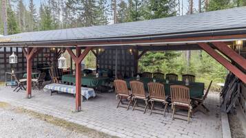Ein überdachter Außenbereich mit Tischen, Stühlen, einem Grill und Blick auf Bäume.