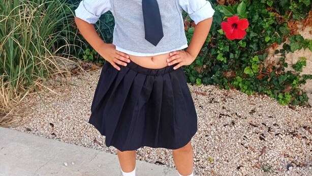 Niño con uniforme escolar de pie al aire libre, con follaje y una flor roja de fondo.