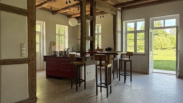 Een keuken met een houten plafond, een rode aanrecht en uitzicht op een groene tuin door grote ramen.