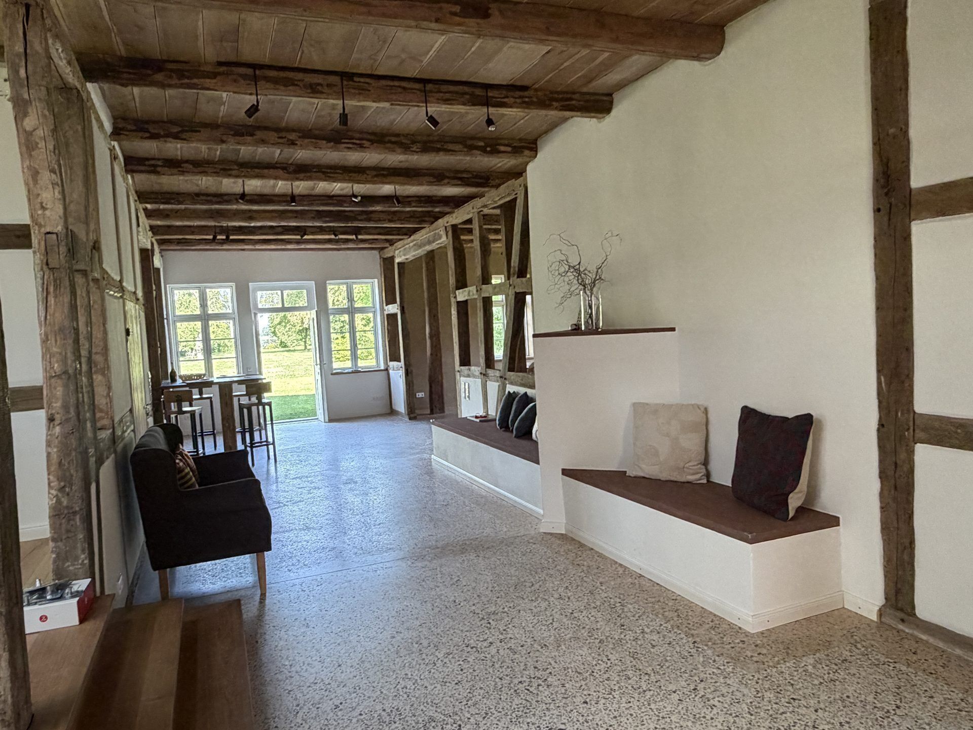 A spacious room with a high ceiling, wooden beams, a seating area with cushions, and large windows allowing natural light.