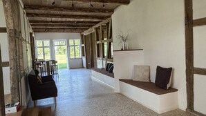 Een ruime kamer met een hoge nok, houten balken, een zithoek met kussens en grote ramen voor natuurlijk licht.