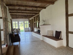 Una habitación amplia con techo alto, vigas de madera, zona de estar con cojines y ventanas grandes que dejan entrar luz natural.