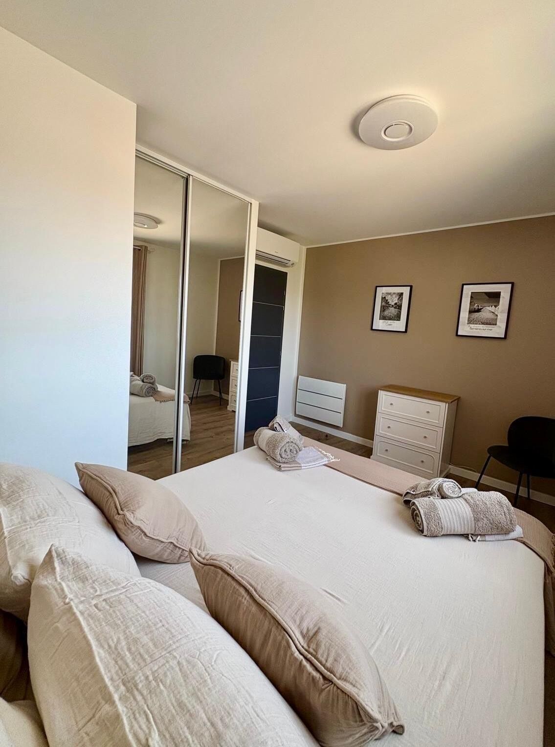 Un dormitorio con cama, almohadas, cómoda, silla, espejo y dos cuadros en la pared.