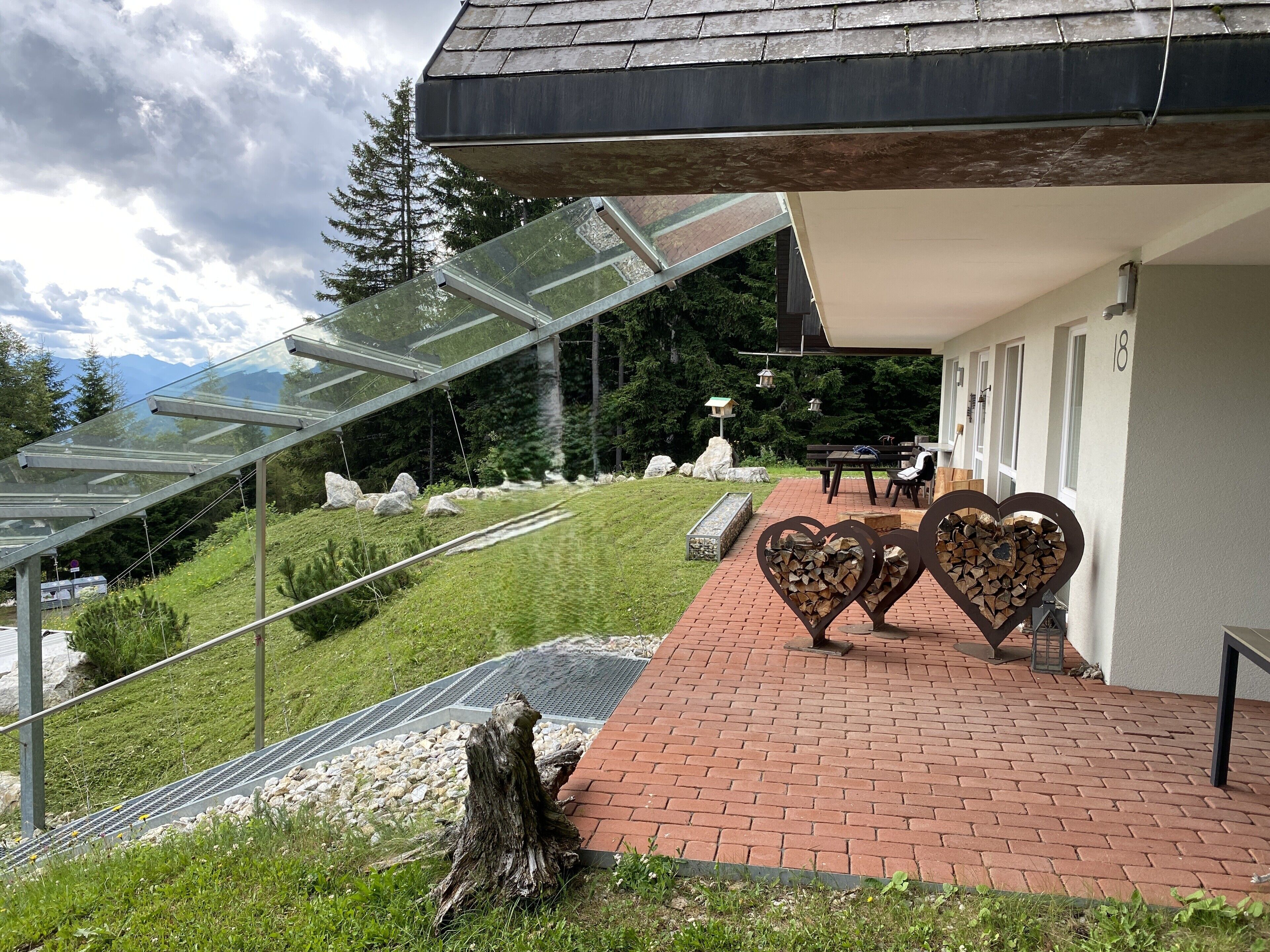 A building with a glass-roofed staircase, a brick pathway, heart-shaped firewood sculptures, and a view of a mountainous landscape.