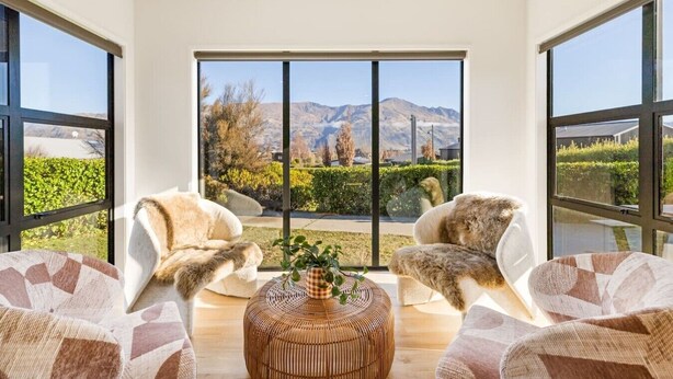 A modern living room with large windows, two fur-covered chairs, and a woven coffee table. - Modern Wanaka Home | Mountain Views | Sleeps 6 (Wānaka)