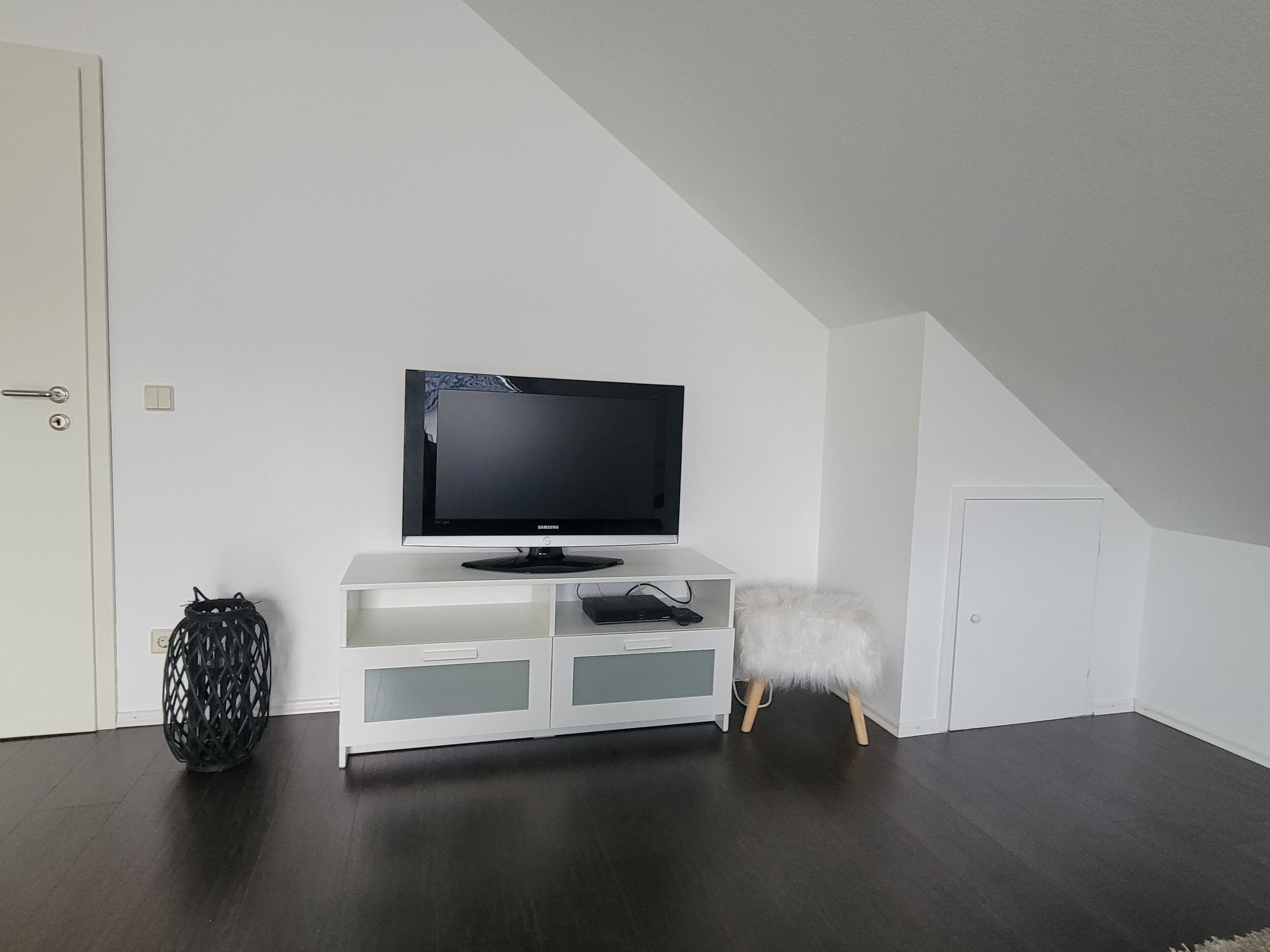 A white entertainment unit with a flat-screen TV, a decorative basket, and a fluffy white stool.