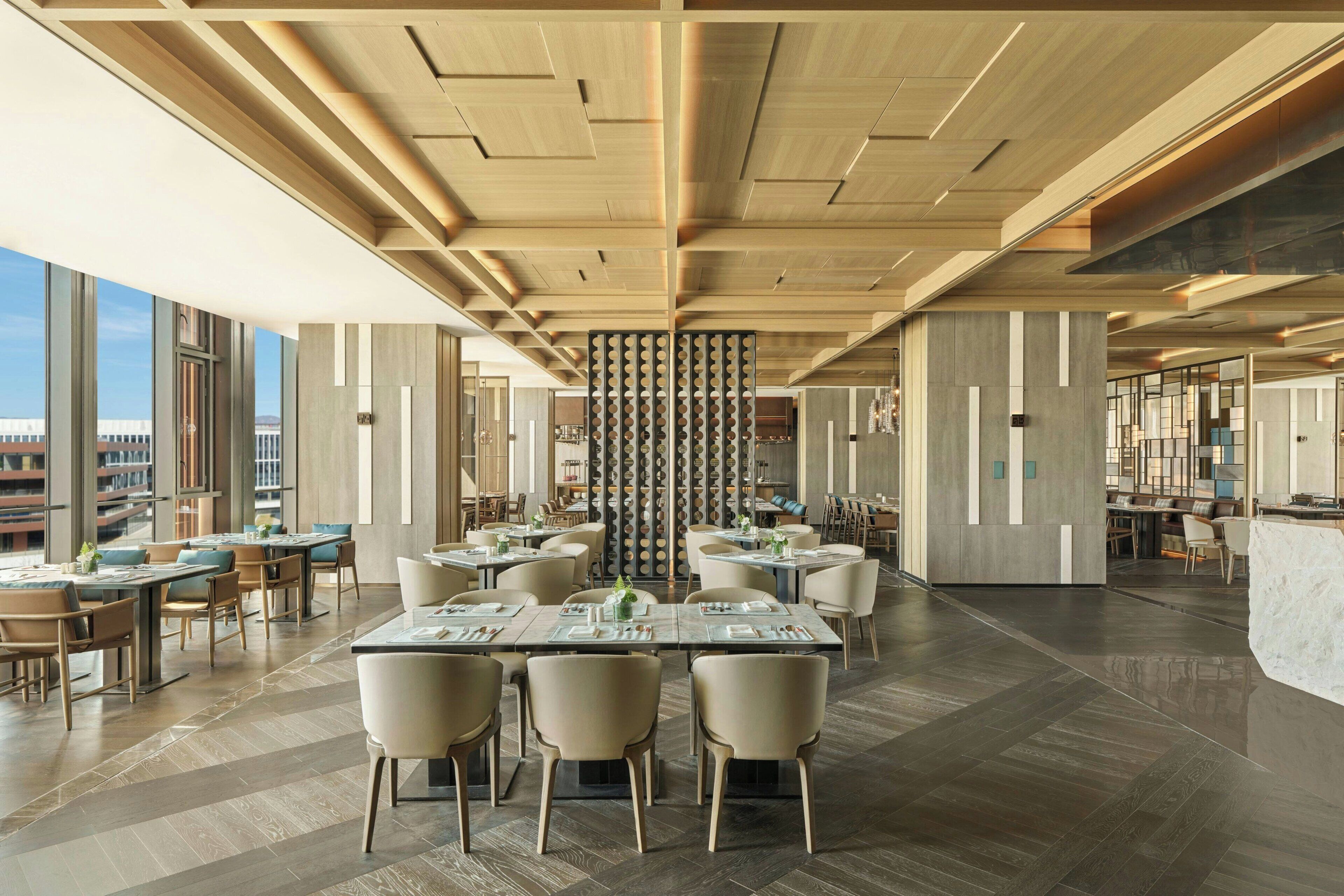 Un restaurante moderno con techos altos, paneles de madera y una columna central con aberturas circulares. Las mesas están dispuestas para comer y el espacio está bien iluminado con luz natural.