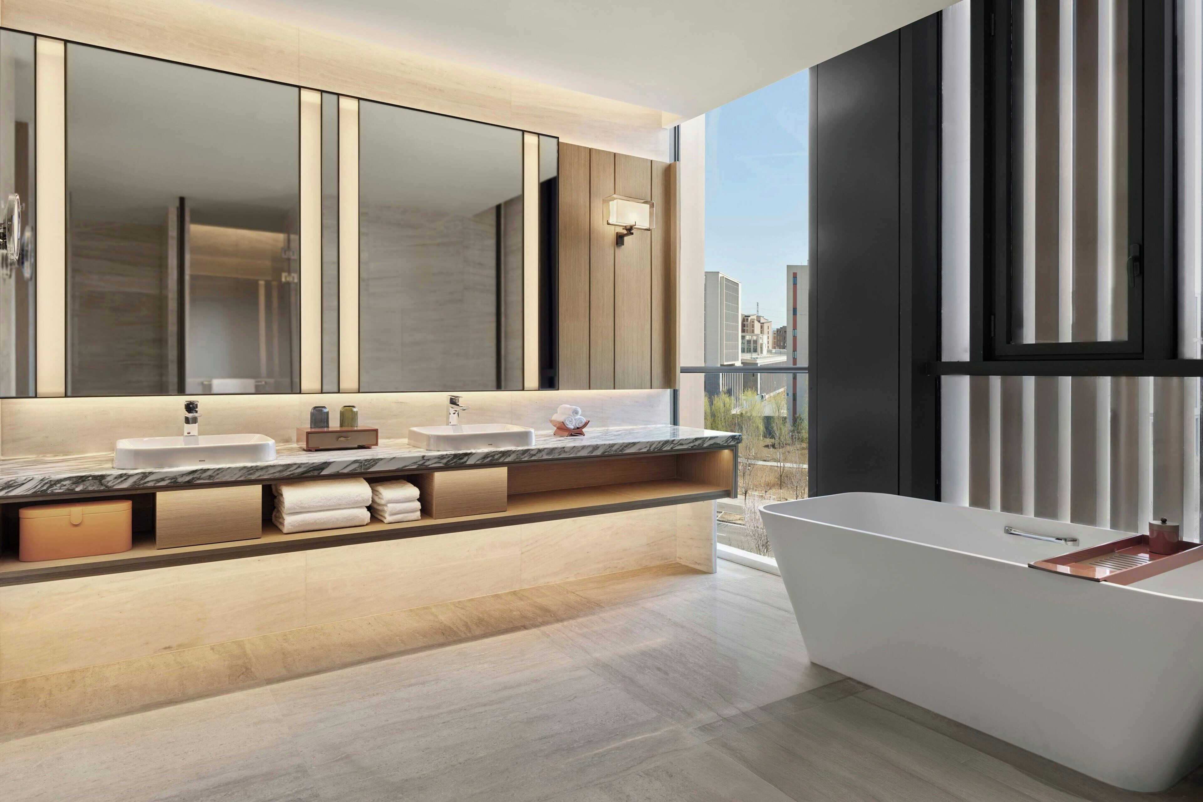 A modern bathroom with a large mirror, two sinks, a bathtub, and a view of the city.