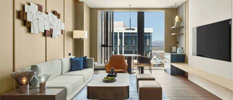 Habitación de hotel moderna con sofá, sillones, una mesita de centro, televisor y vistas a la ciudad.