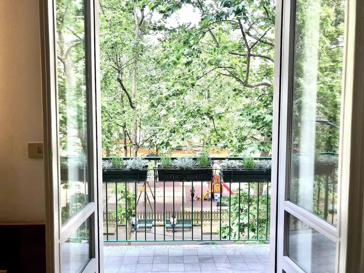 A view through a glass door leading to a balcony with a metal railing, a tiled floor, and a green fence in the background.