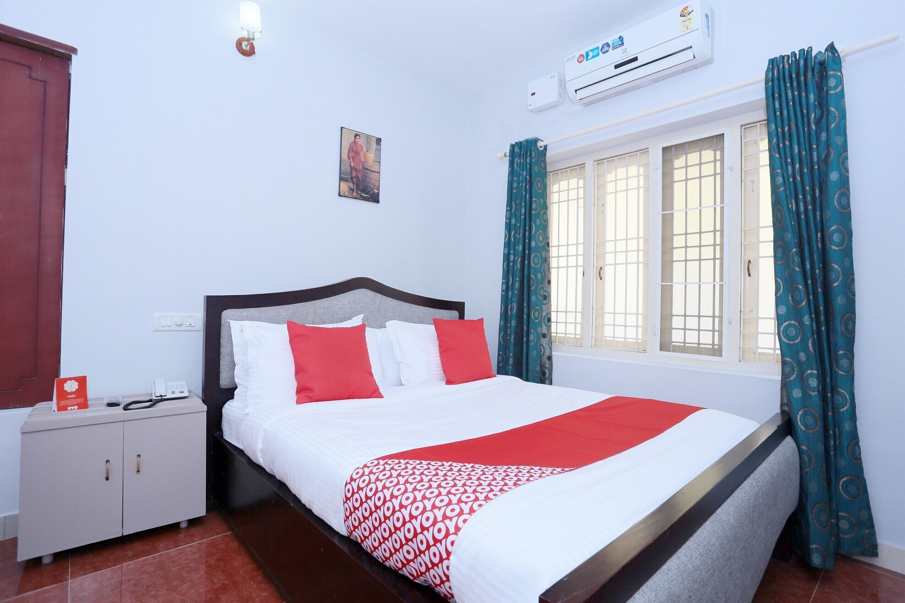 A neatly made bed with a patterned blanket, a bedside table with a phone and a card, a wall-mounted air conditioner, and a window with curtains.