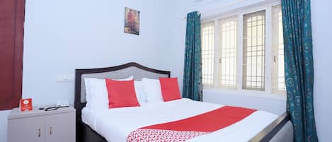 A neatly made bed with a patterned blanket, a bedside table with a phone and a card, a wall-mounted air conditioner, and a window with curtains.