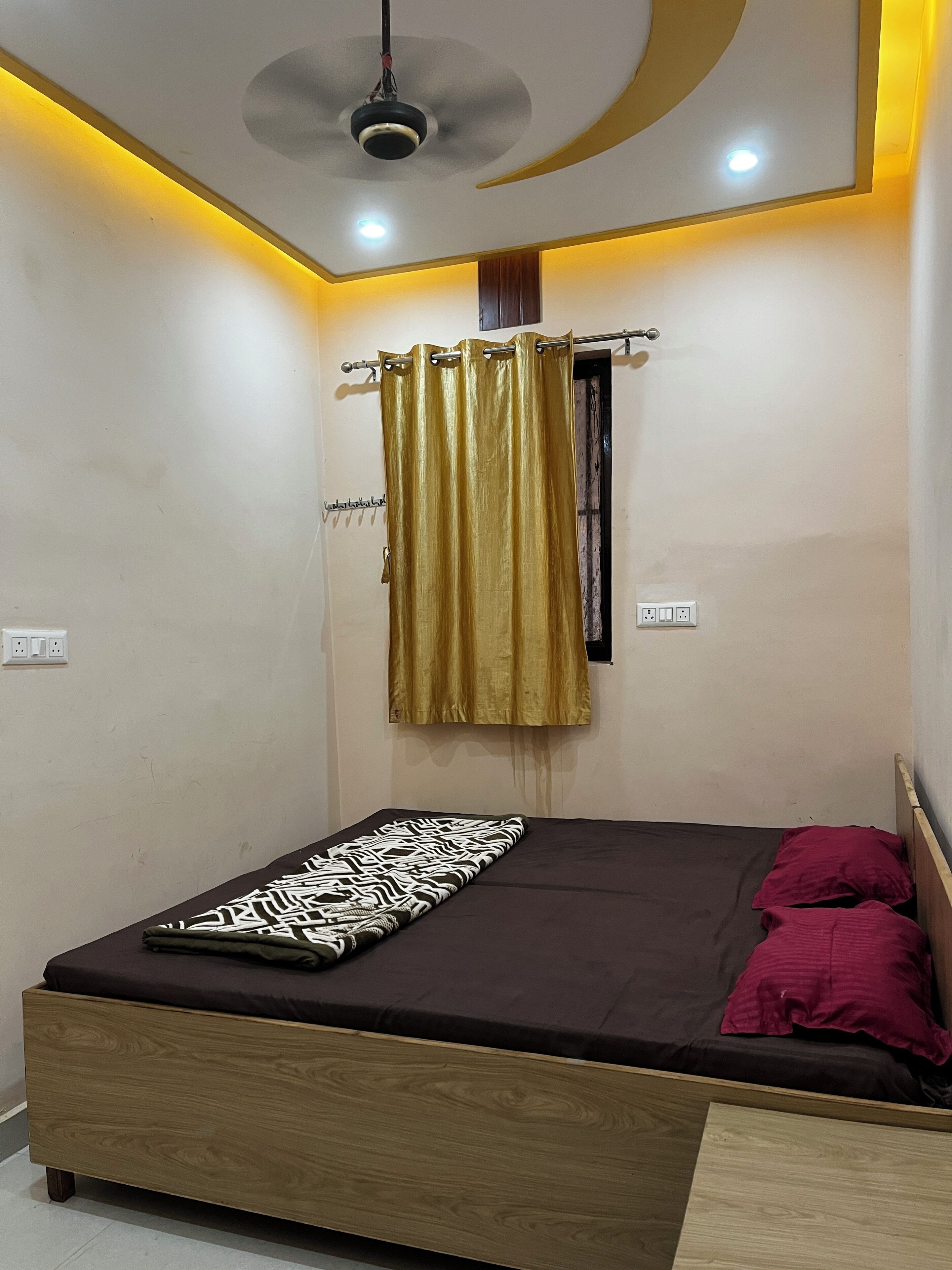 A bedroom with a bed, a ceiling fan, a curtain, and a wall socket.