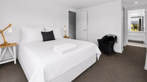 Une chambre d’hôtel moderne avec un grand lit, du linge de lit blanc, une table de chevet en bois avec une lampe, une chaise noire et une vue sur l’extérieur par une porte ouverte.