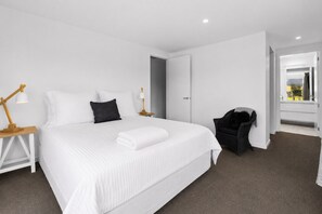 Ein modernes Hotelzimmer mit einem großen Bett, weißer Bettwäsche, einem Nachttisch aus Holz mit einer Lampe, einem schwarzen Sessel und Blick ins Freie durch eine offene Tür.
